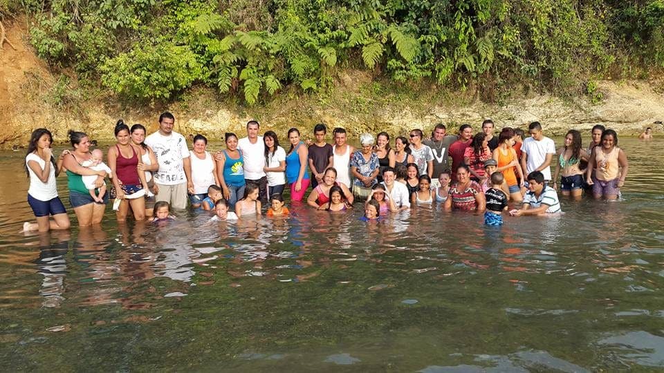 Decenas de personas de la congregación, adultos y niños, posan dentro del río con la vegetación de la ribera detrás