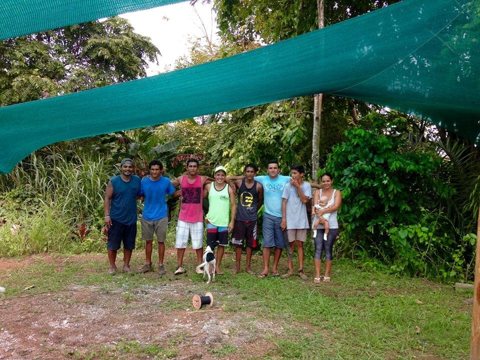 Un grupo de jóvenes y adultos posa bajo una malla de sombra verde, rodeado de vegetación