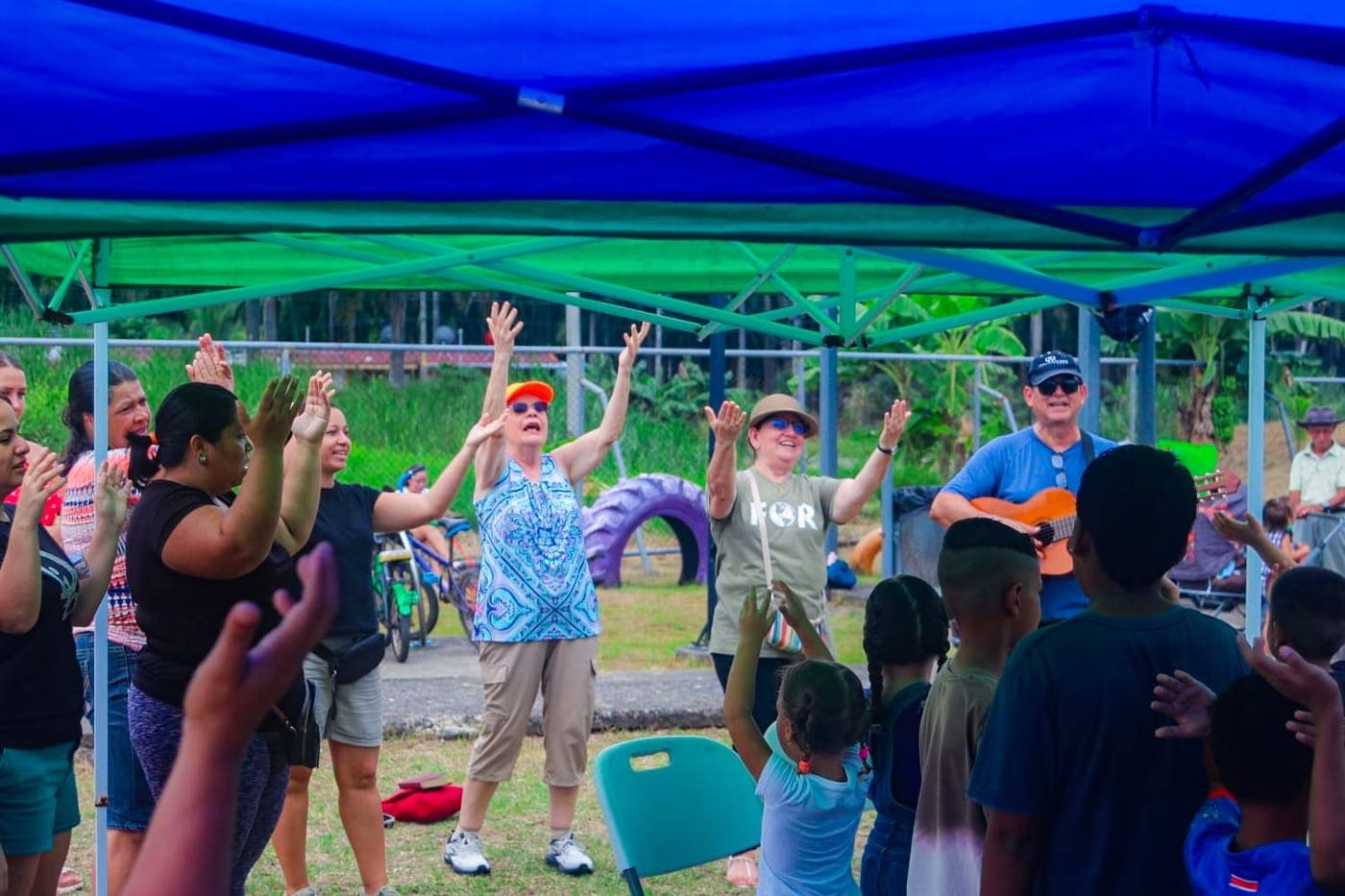 Niños y adultos con las manos alzadas durante una actividad al aire libre, bajo un toldo azul