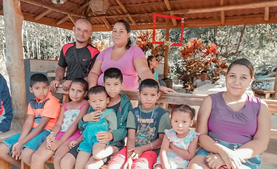 Personas de la comunidad compartiendo alrededor de mesas, bajo el techo del salón decorado con flores