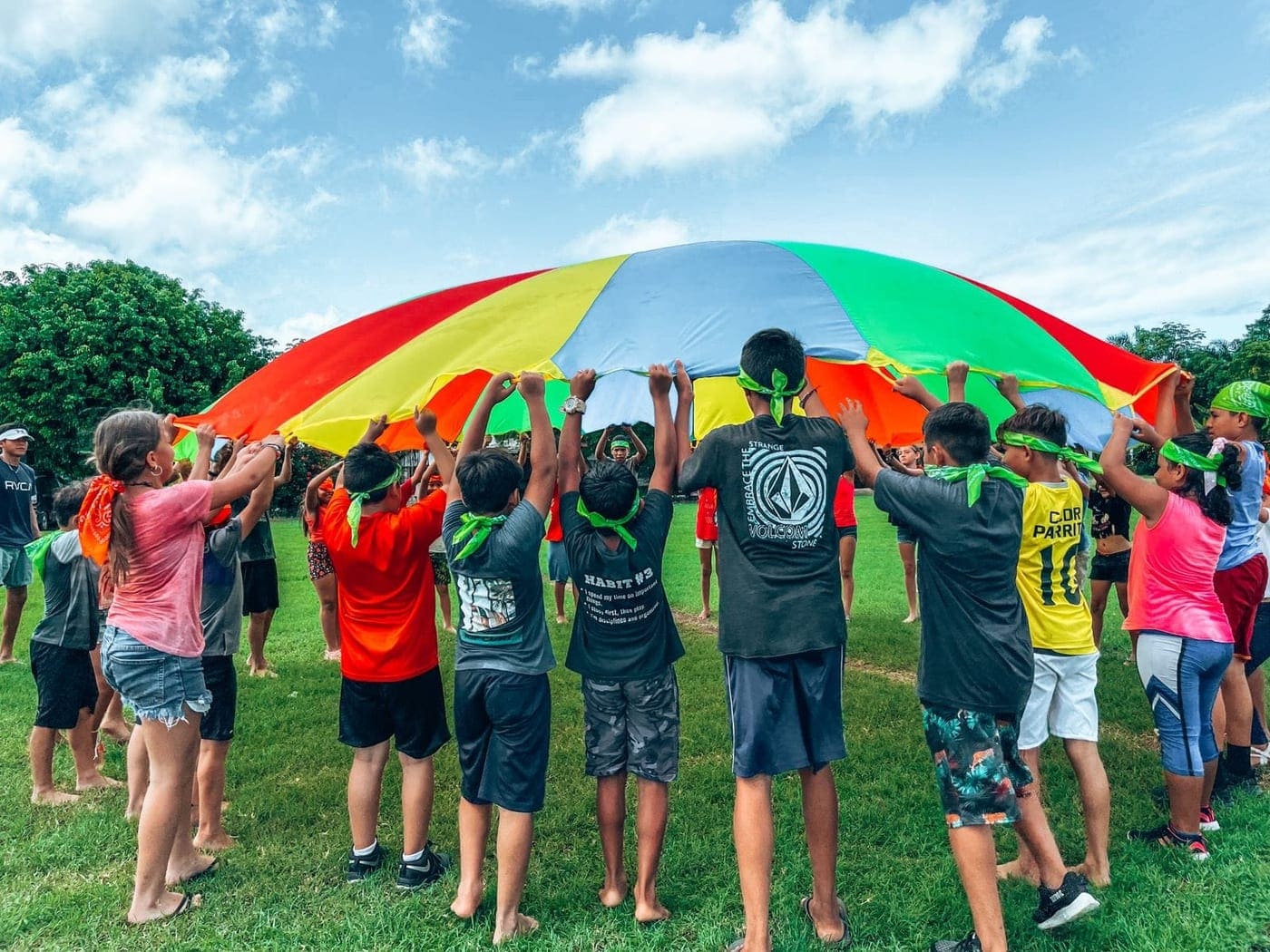 Niños del club levantan juntos un paracaídas de colores sobre el césped, bajo un cielo azul con nubes