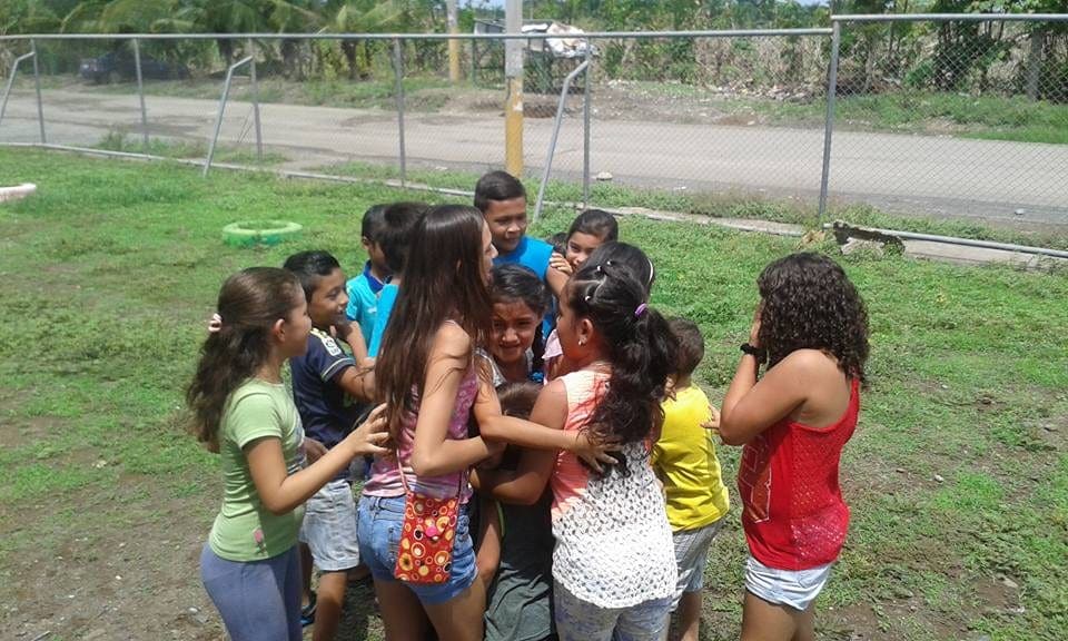Niñas y niños forman una ronda tomados de las manos sobre el césped, junto a la malla de la cancha