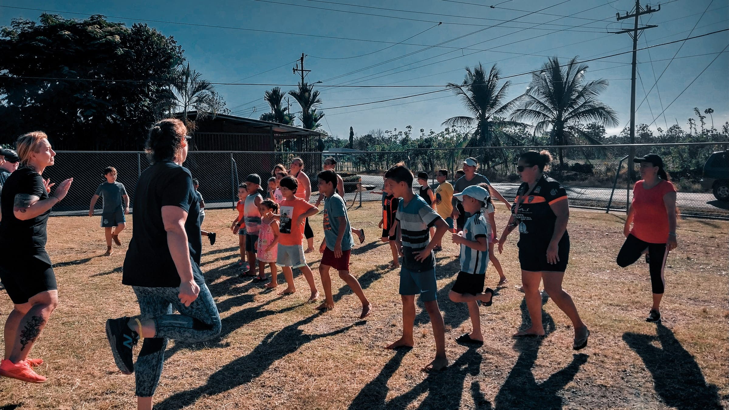 Niños y adultos del club Valores Kids caminando y estirando juntos sobre la cancha de tierra, con palmeras y cielo abierto de Parrita al fondo