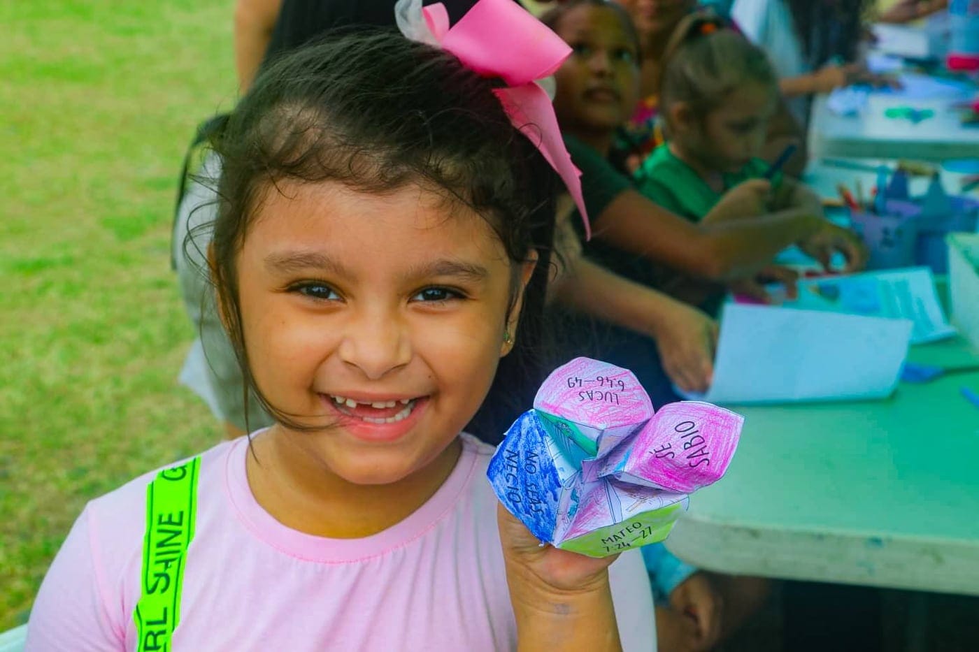 Una niña sonríe sosteniendo la manualidad de papel que acaba de armar, con otros niños del club trabajando en la mesa detrás de ella